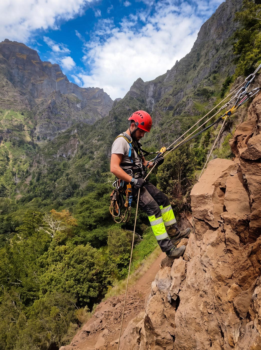 Técnico em rapel numa escarpa da Madeira, com vale verde ao fundo